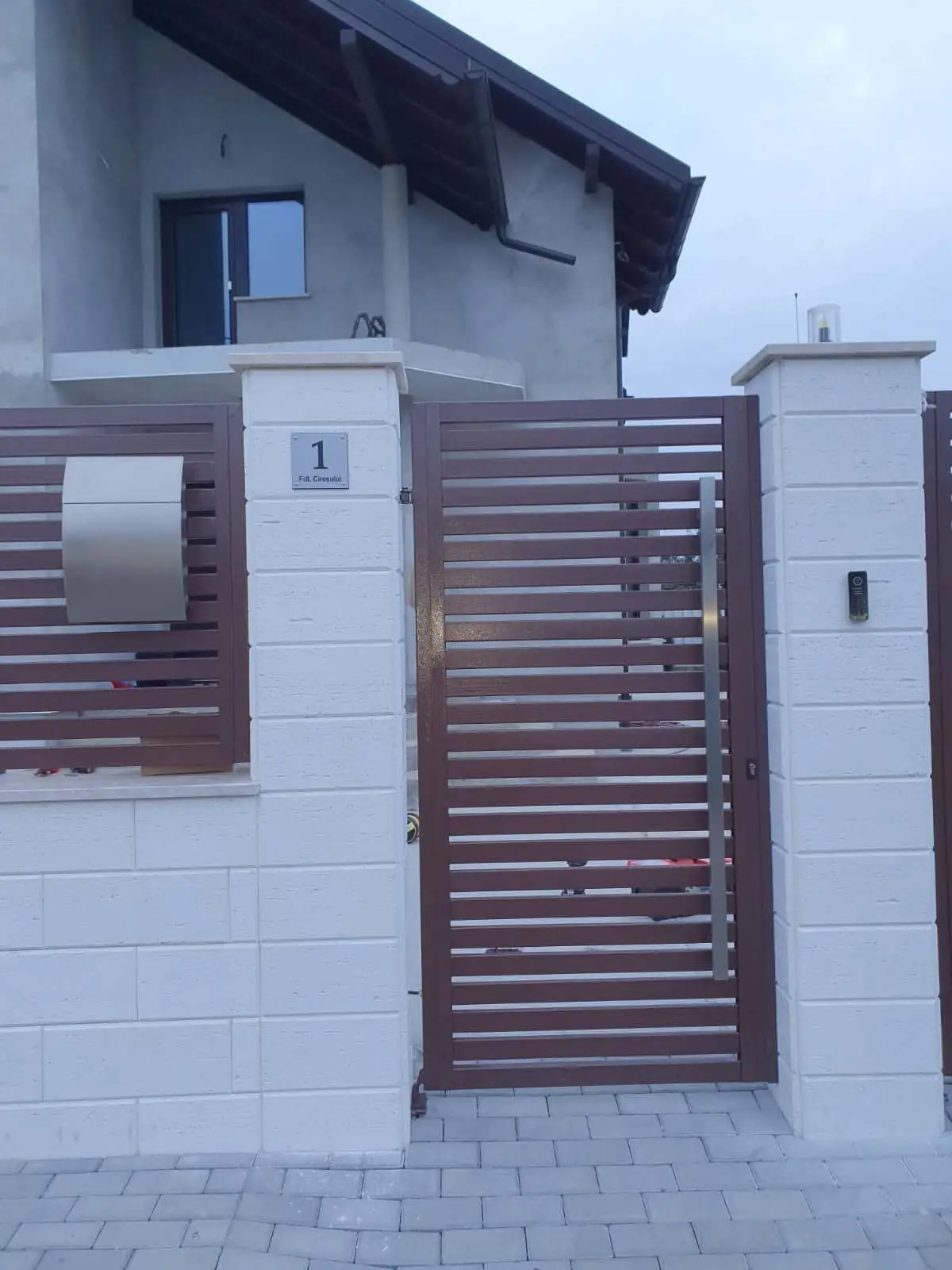 Poartă pietonală din metal cu lamele maro, montată între stâlpi albi
